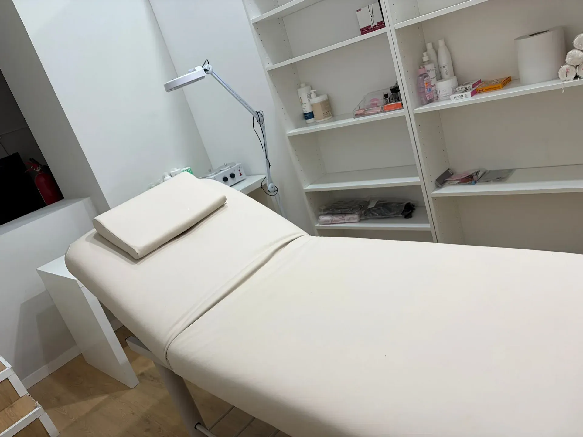 Sala de tratamientos estéticos con mesa de masajes beige, estanterías y lámpara de trabajo.