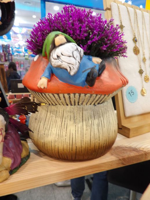 Figura de gnomo sonriente en una maceta con forma de seta y flores moradas en el estante de una tienda.