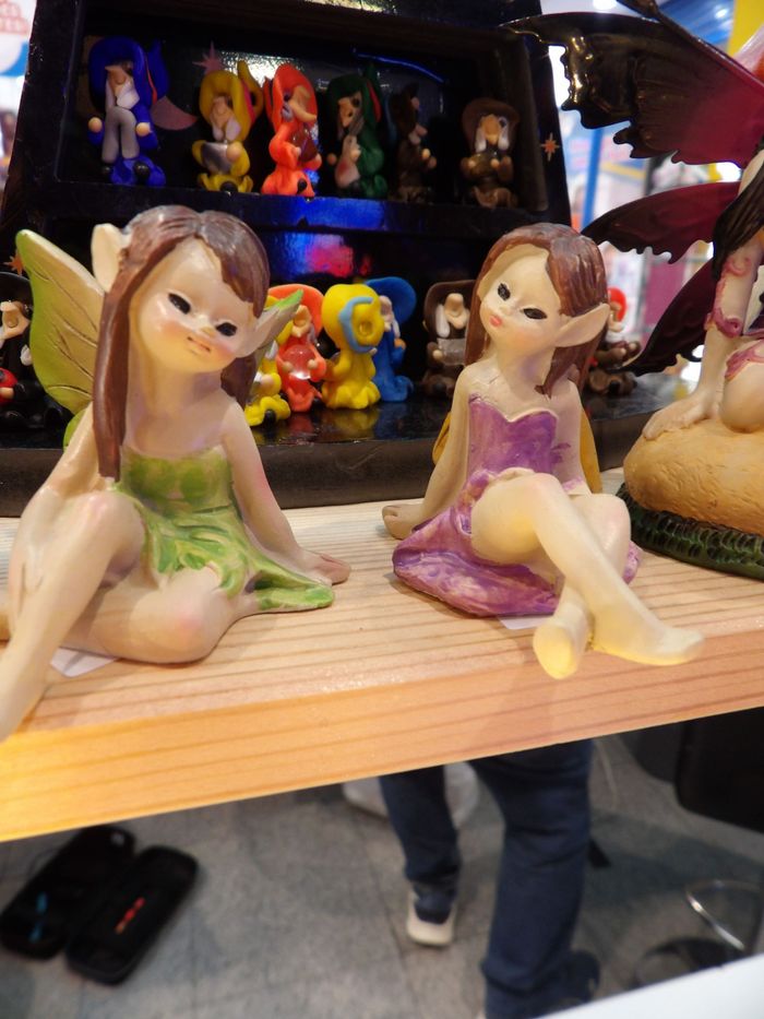 Dos pequeñas figuritas de hadas sentadas sobre una repisa de madera, con coloridos juguetes al fondo.