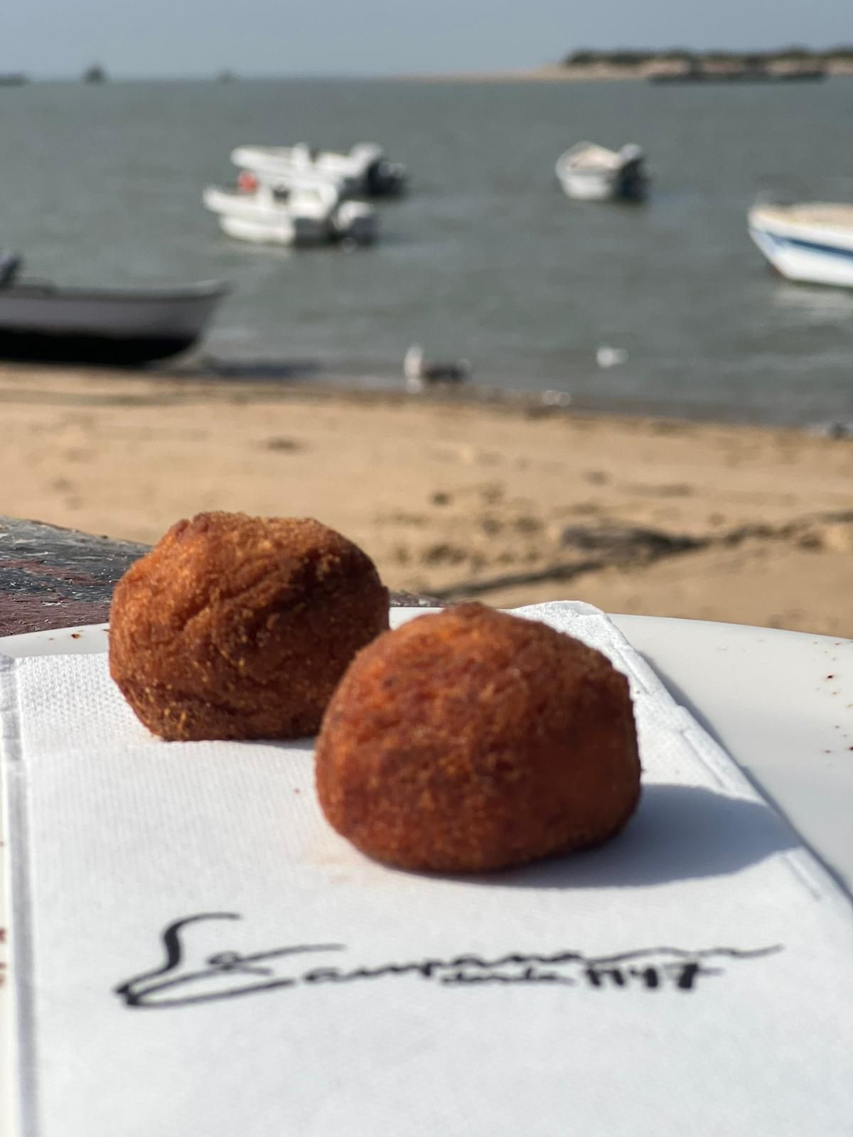 Dos croquetas fritas sobre una servilleta con una playa y barcos de fondo.