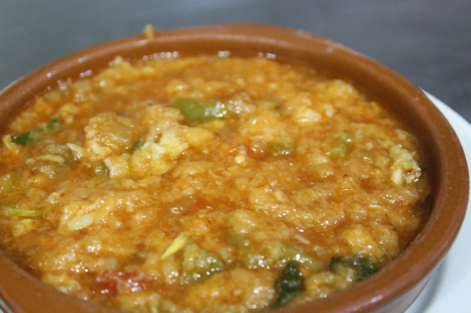 Primer plano de un recipiente rústico, redondo y marrón lleno de un guiso de color naranja rojizo con trozos visibles de color verde y blanco.