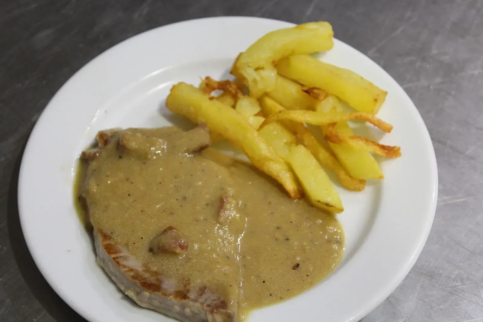 Bistec cubierto de salsa, junto a una pila de papas fritas en un plato blanco.