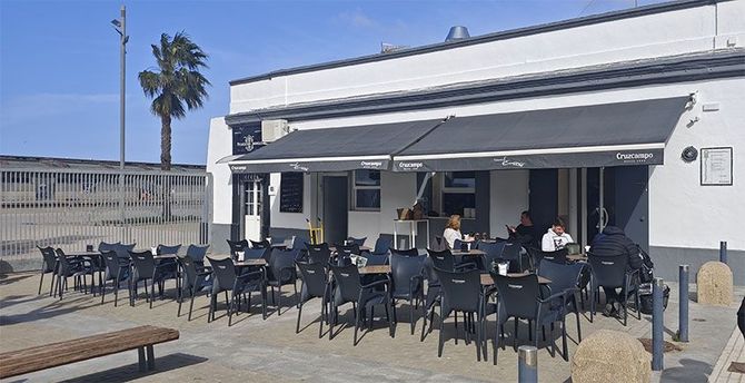 Cafetería al aire libre con mesas y sillas negras, bajo un toldo negro. Gente sentada en las mesas.