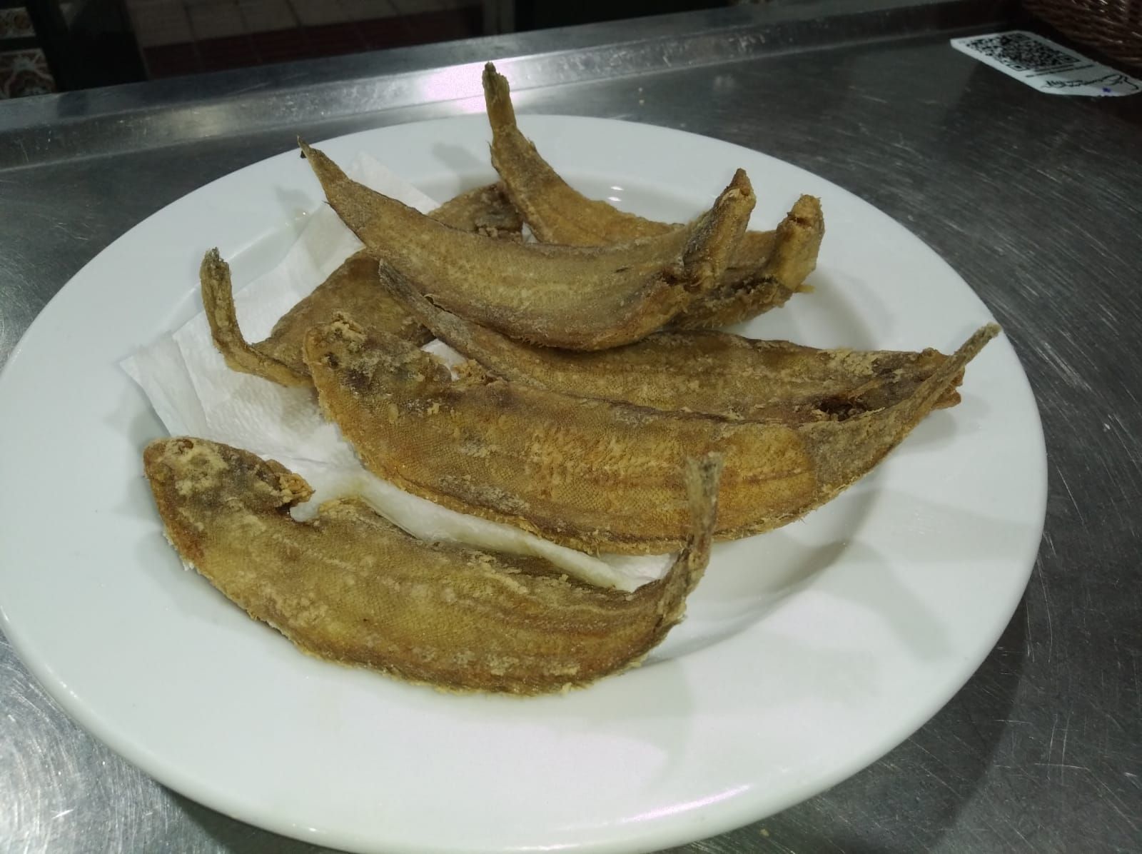 Pescado frito, dorado, en un plato blanco, servido sobre una superficie metálica.