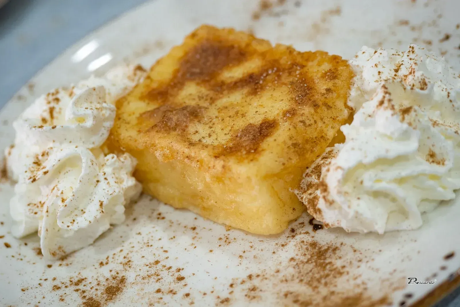 Budín de pan frito, cubierto con canela, servido con crema batida en un plato blanco.