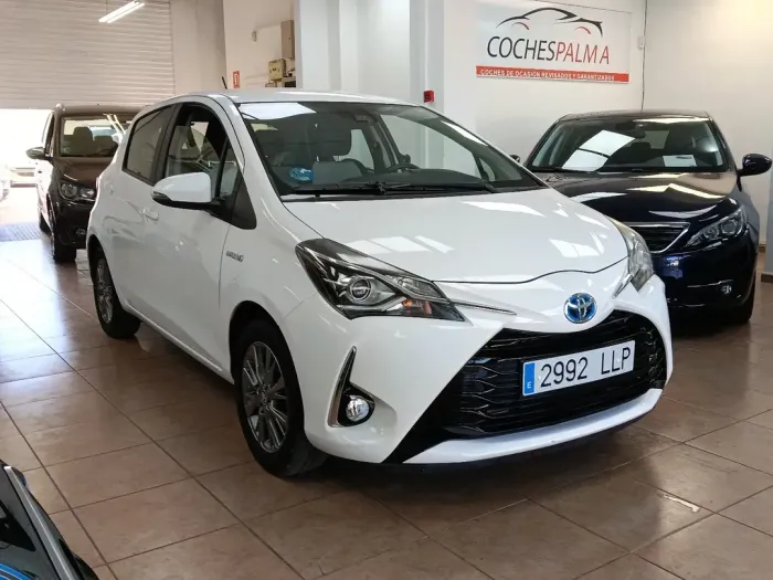 Un Toyota hatchback blanco en una sala de exposición de automóviles con otros vehículos al fondo.