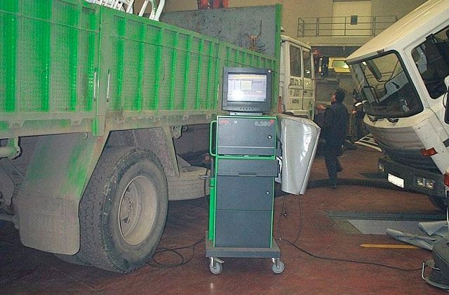 Camión averiado y furgoneta volcada en un taller, con un pequeño monitor CRT sobre un carrito en primer plano.