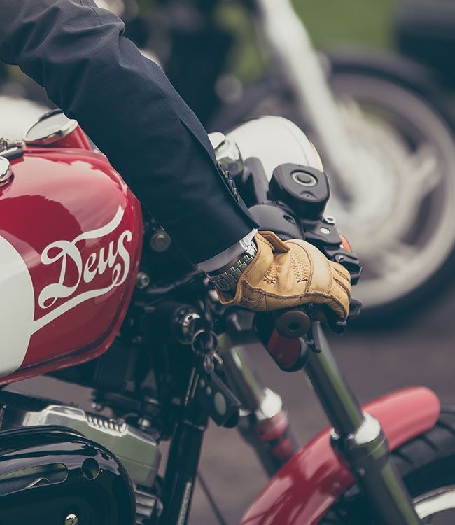 Main gantée de cuir beige sur le guidon d'une moto, près d'un réservoir d'essence rouge et blanc orné du logo « Deus ».