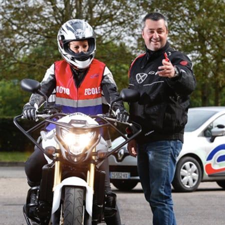 Élève motard sur sa moto, moniteur pointant du doigt. Scène extérieure avec une voiture en arrière-plan.