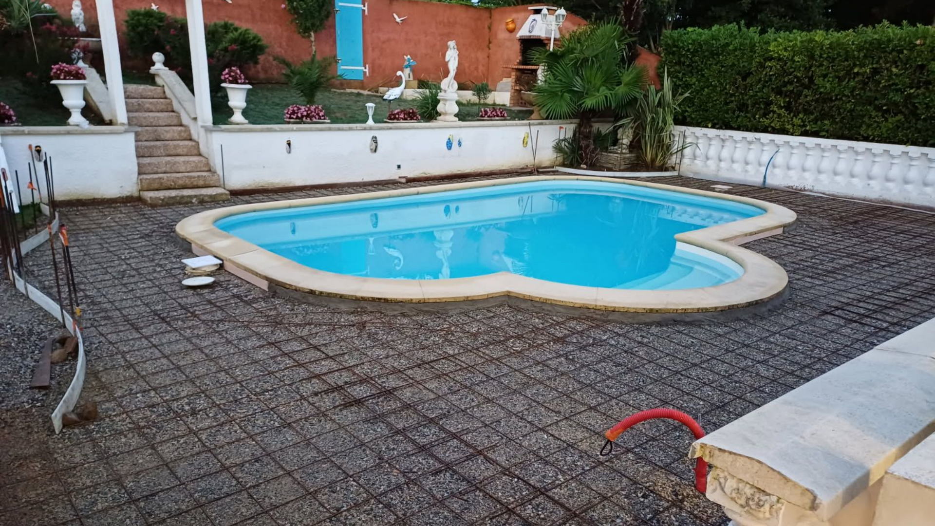 Une piscine en forme de cœur entourée de grillage, avec un patio, des escaliers et des statues visibles.