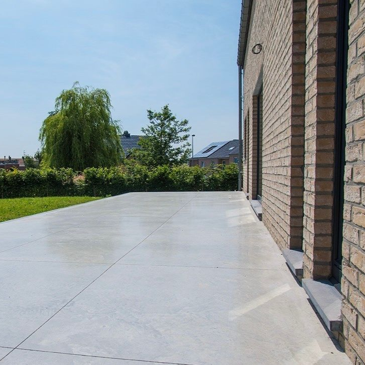 Un patio en béton gris à côté d'une maison en briques, avec de l'herbe et des arbres en arrière-plan.