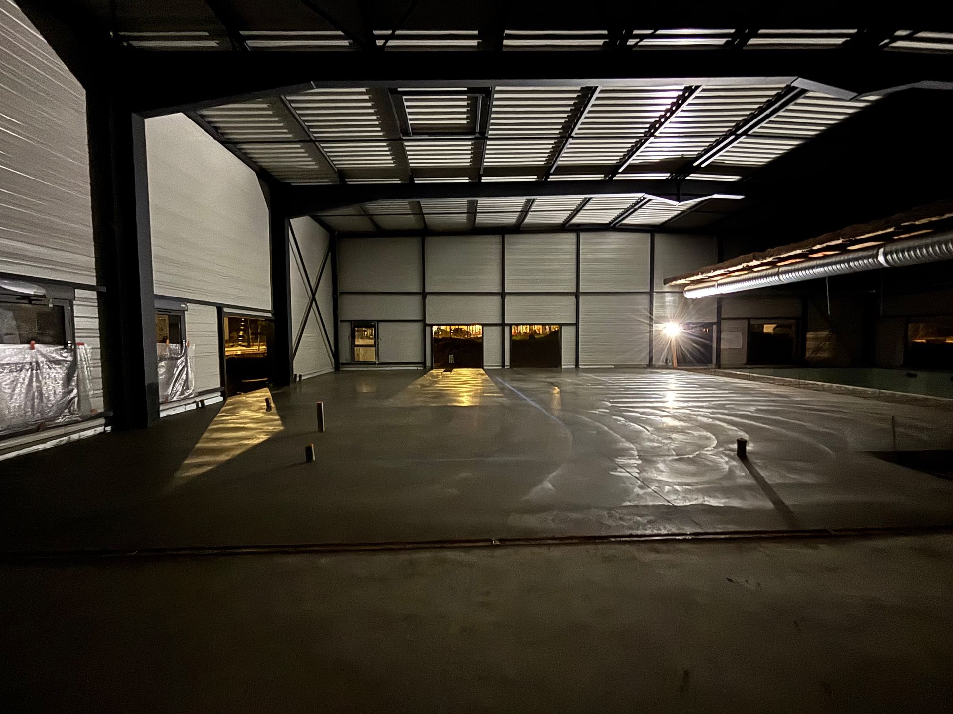 Intérieur d'un entrepôt métallique vide la nuit, sol en béton fraîchement coulé reflétant les faibles lumières du plafond.