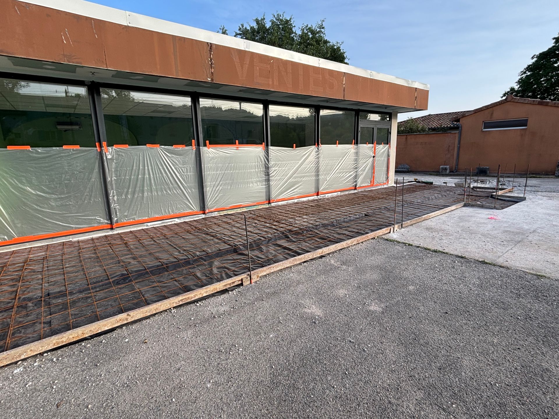 Fondations en béton avec barres d'armature devant un bâtiment aux grandes fenêtres.