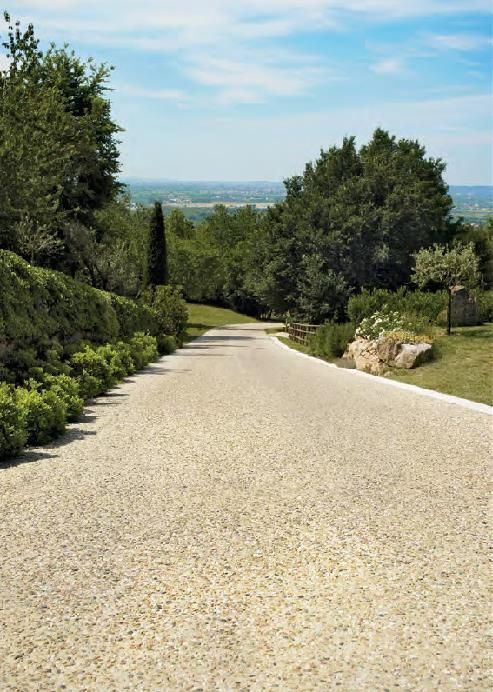 Allée de graviers bordée de buissons verts et d'arbres, menant à une vue lointaine sur une vallée.
