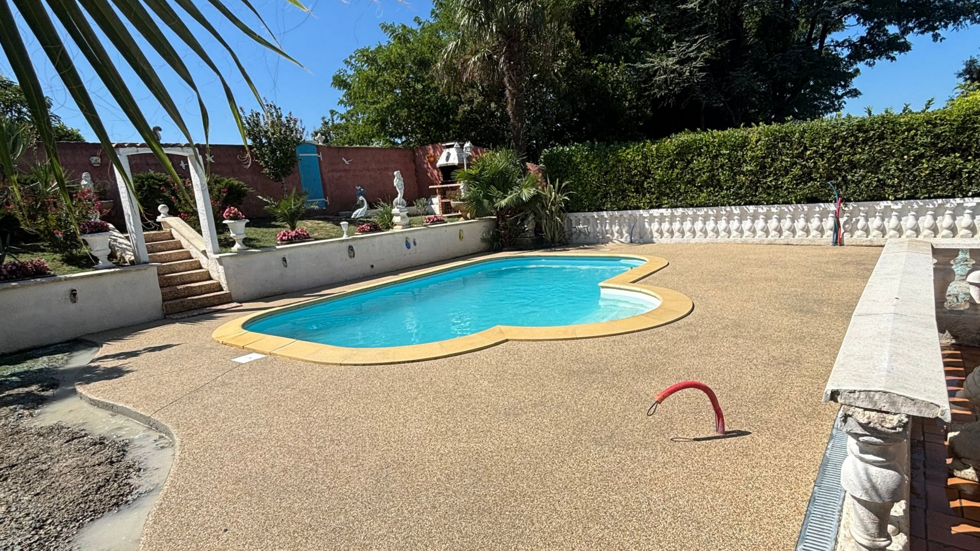 Piscine avec bordure de galets beiges, eau bleue, clôture blanche et arbustes.