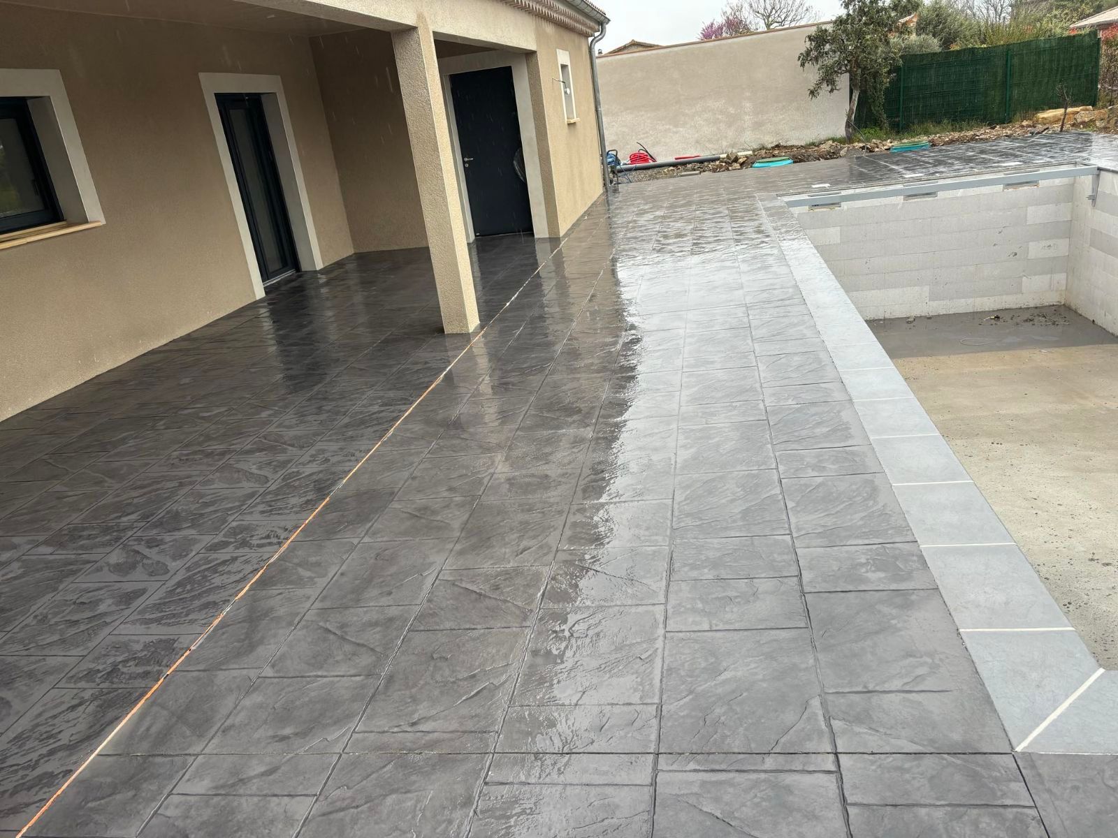 Patio carrelé gris humide à côté d'une piscine en construction, rattachée à un bâtiment beige.