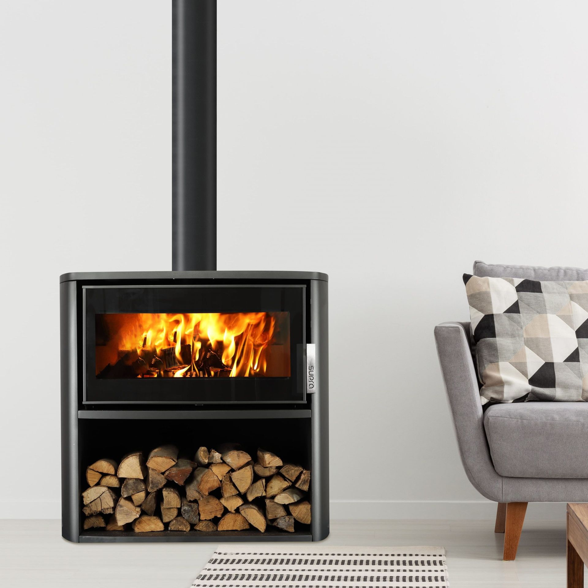Poêle à bois noir moderne avec un feu qui brûle, bûches stockées en dessous, à côté d'un canapé gris avec des coussins à motifs.