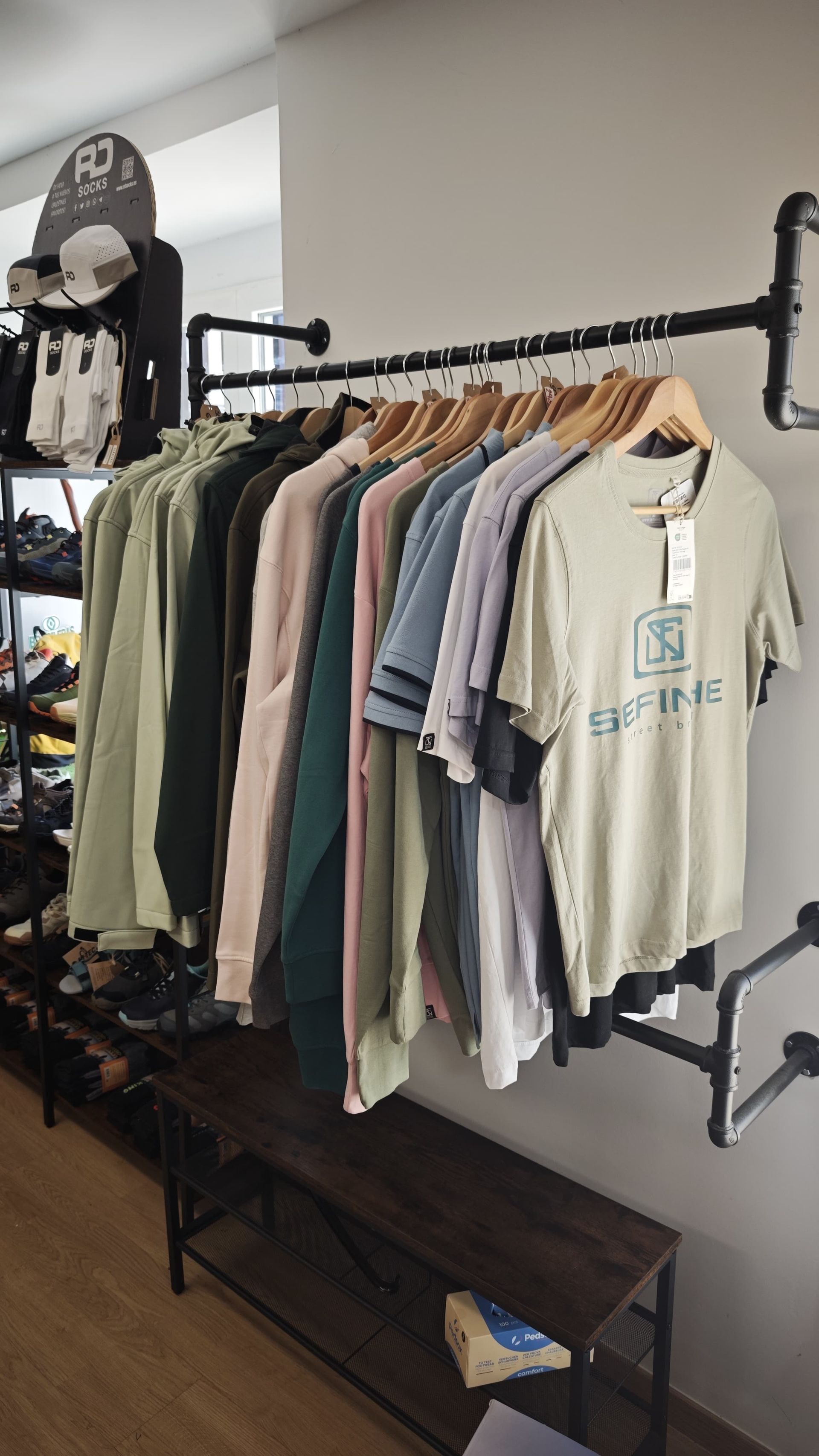 Percheros con camisetas variadas en una tienda de ropa, junto a una estantería negra y un expositor de pared