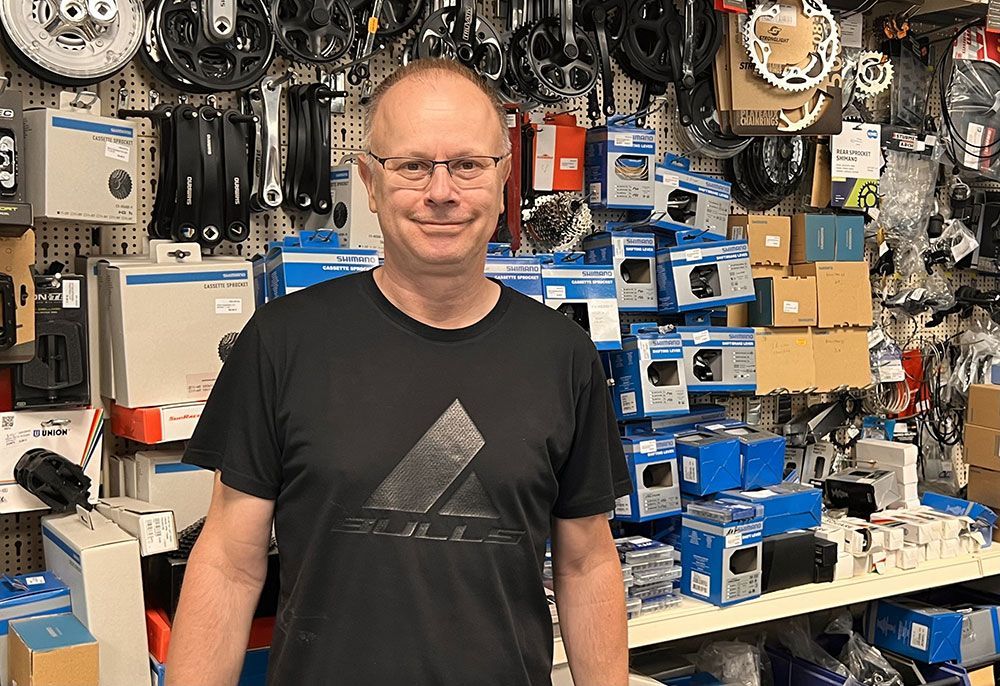 Ein Mann im schwarzen Hemd lächelt in einem Fahrradladen. Er ist umgeben von Fahrradteilen.