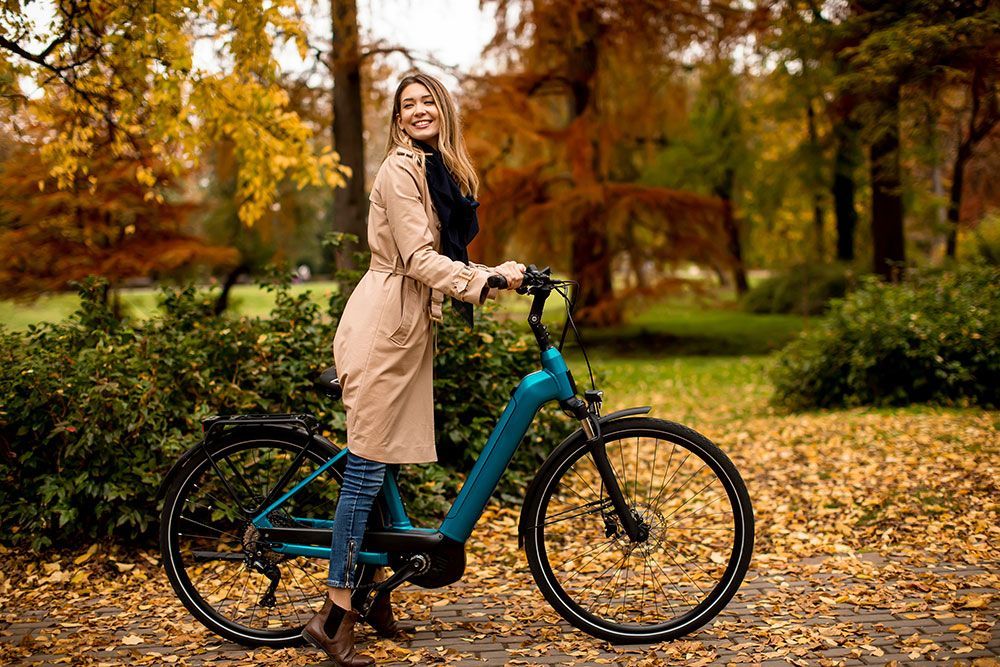 Eine Frau in einem beigen Mantel und Jeans lächelt neben einem türkisfarbenen Elektrofahrrad in einem Park mit Herbstlaub.