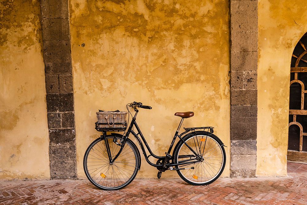 Schwarzes Fahrrad mit Weidenkorb, abgestellt an einer strukturierten gelben Mauer mit Steinsäulen und Bogenfenster.