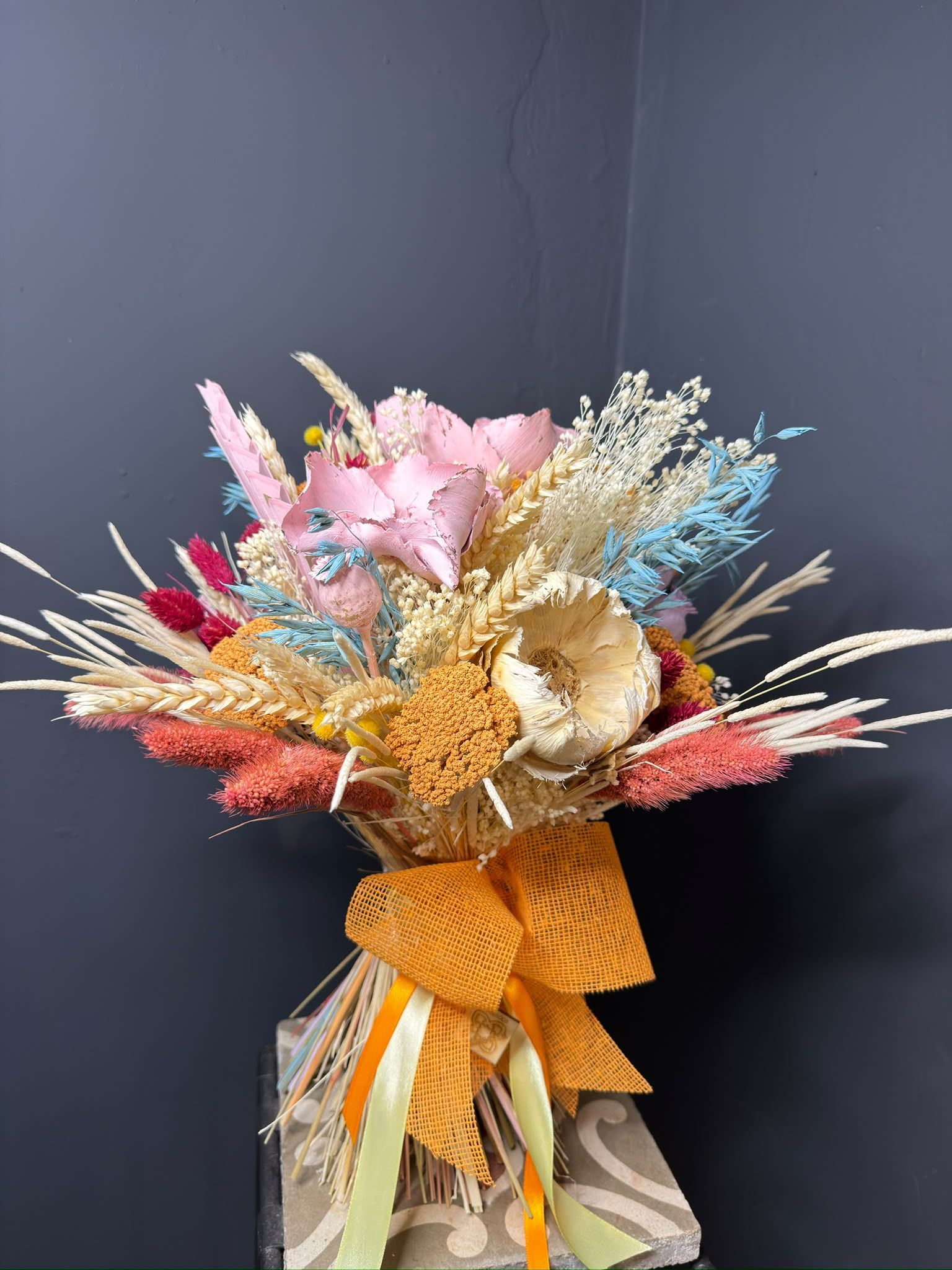 Ramo de flores secas de colores con flores rosas, azules y crema atadas con una cinta naranja sobre un fondo oscuro.