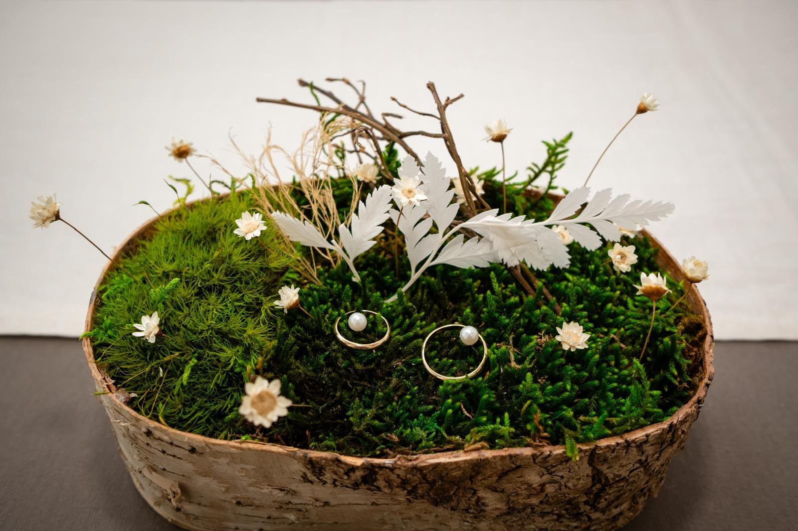 Cesta forrada de musgo con flores blancas y dos anillos de cristal sobre una mesa.