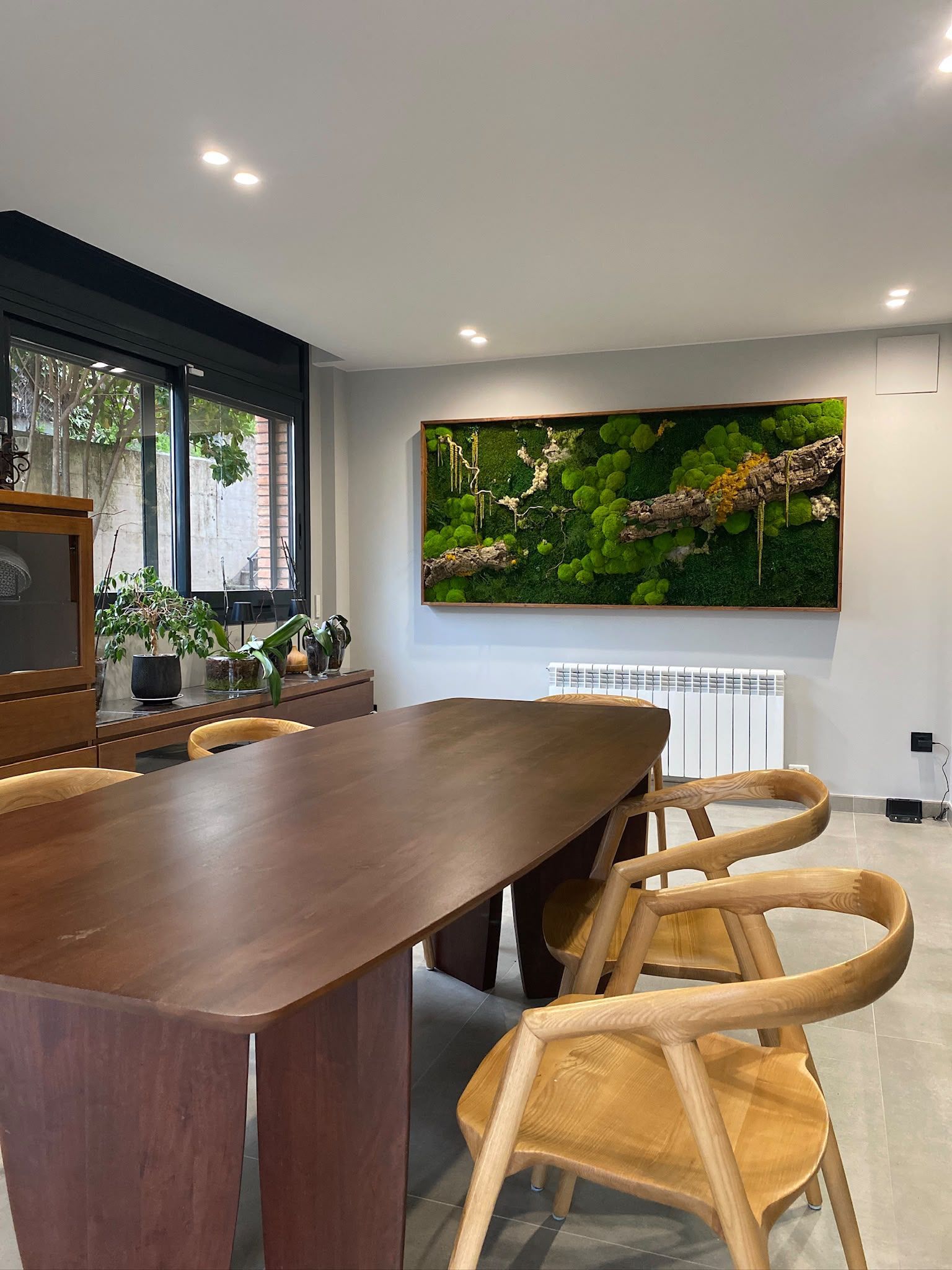 Comedor moderno con mesa y sillas de madera, un gran cuadro en la pared y plantas de interior junto a la ventana.