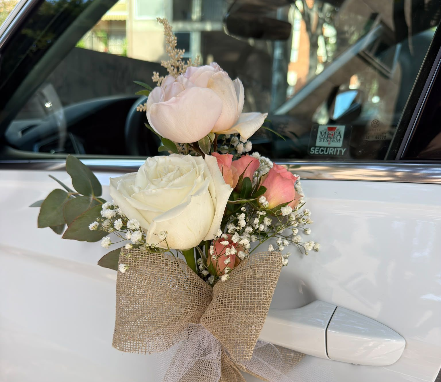 Ramo de flores envuelto en arpillera sujeto a la manija de la puerta de un coche blanco.
