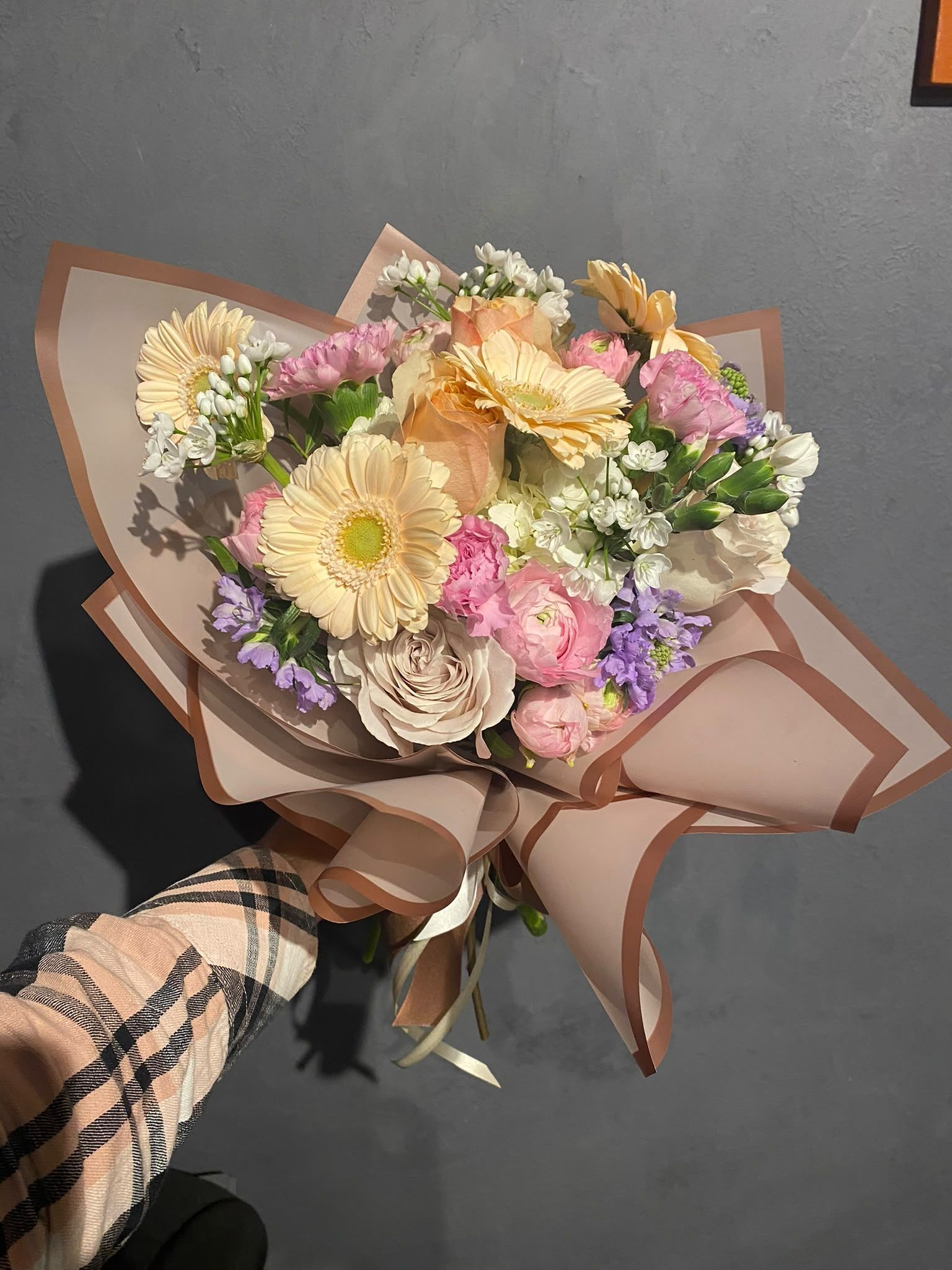 Ramo de flores en tonos pastel envuelto en papel marrón sobre un fondo oscuro.