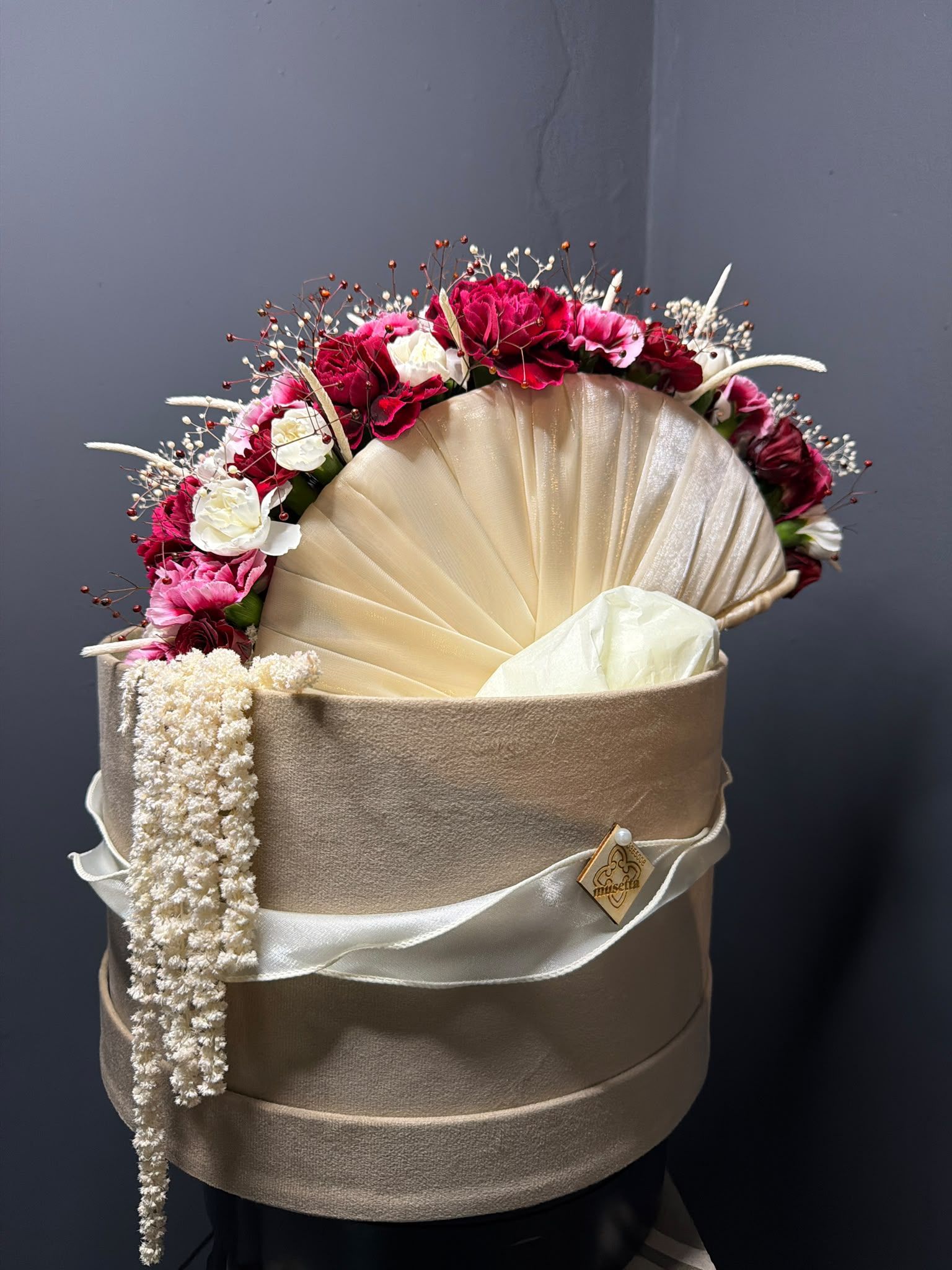 Cesta de regalo decorativa en tonos beige y dorado con flores rosas y blancas sobre un fondo gris.