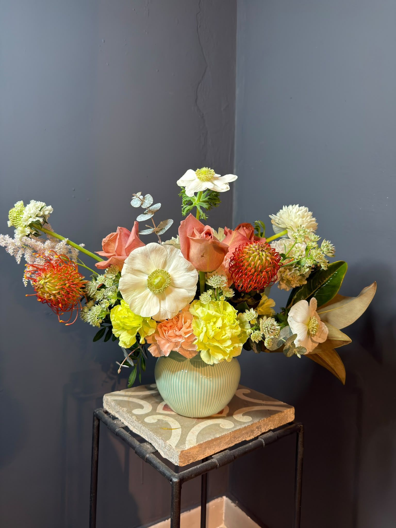Arreglo floral colorido en un jarrón claro sobre una mesa pequeña contra una pared gris.