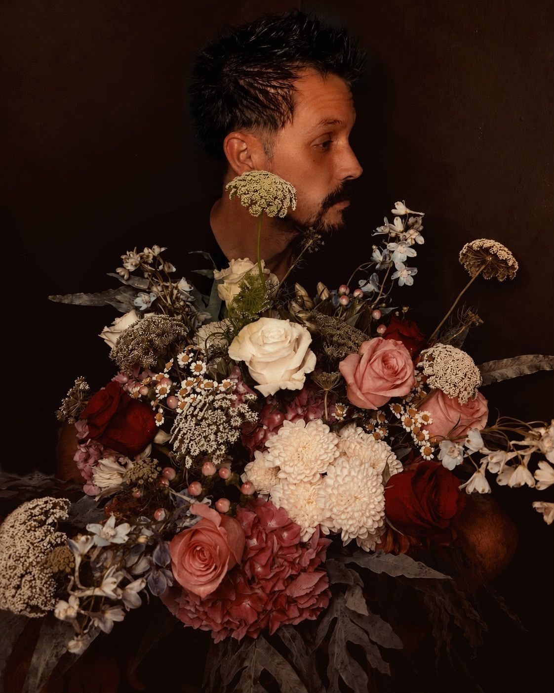 Retrato de una persona de perfil, parcialmente oculta por un exuberante ramo de rosas y flores blancas.
