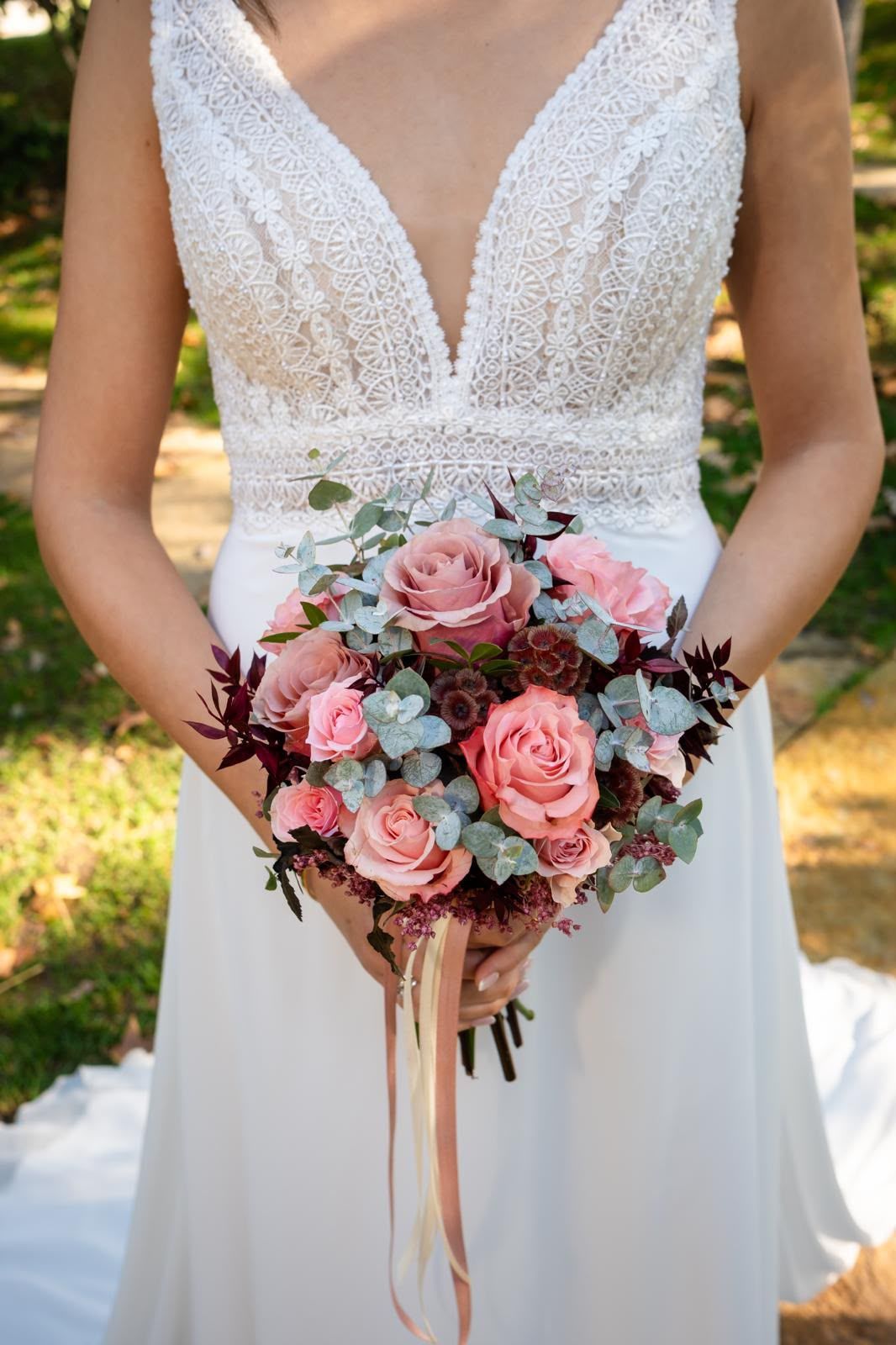 Novia con un vestido de novia de encaje blanco al aire libre, sosteniendo un ramo rosa y burdeos.
