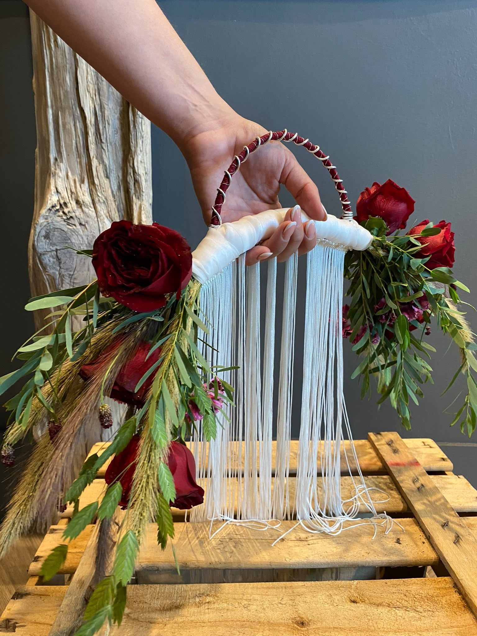 Una mano sostiene una cesta roja con flores y cintas blancas sobre una caja de madera decorada con rosas y follaje.