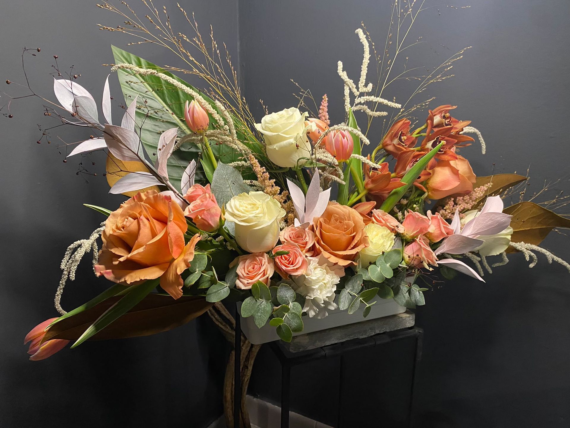 Arreglo floral con flores naranjas, crema y rosas en un jarrón negro sobre un fondo gris.