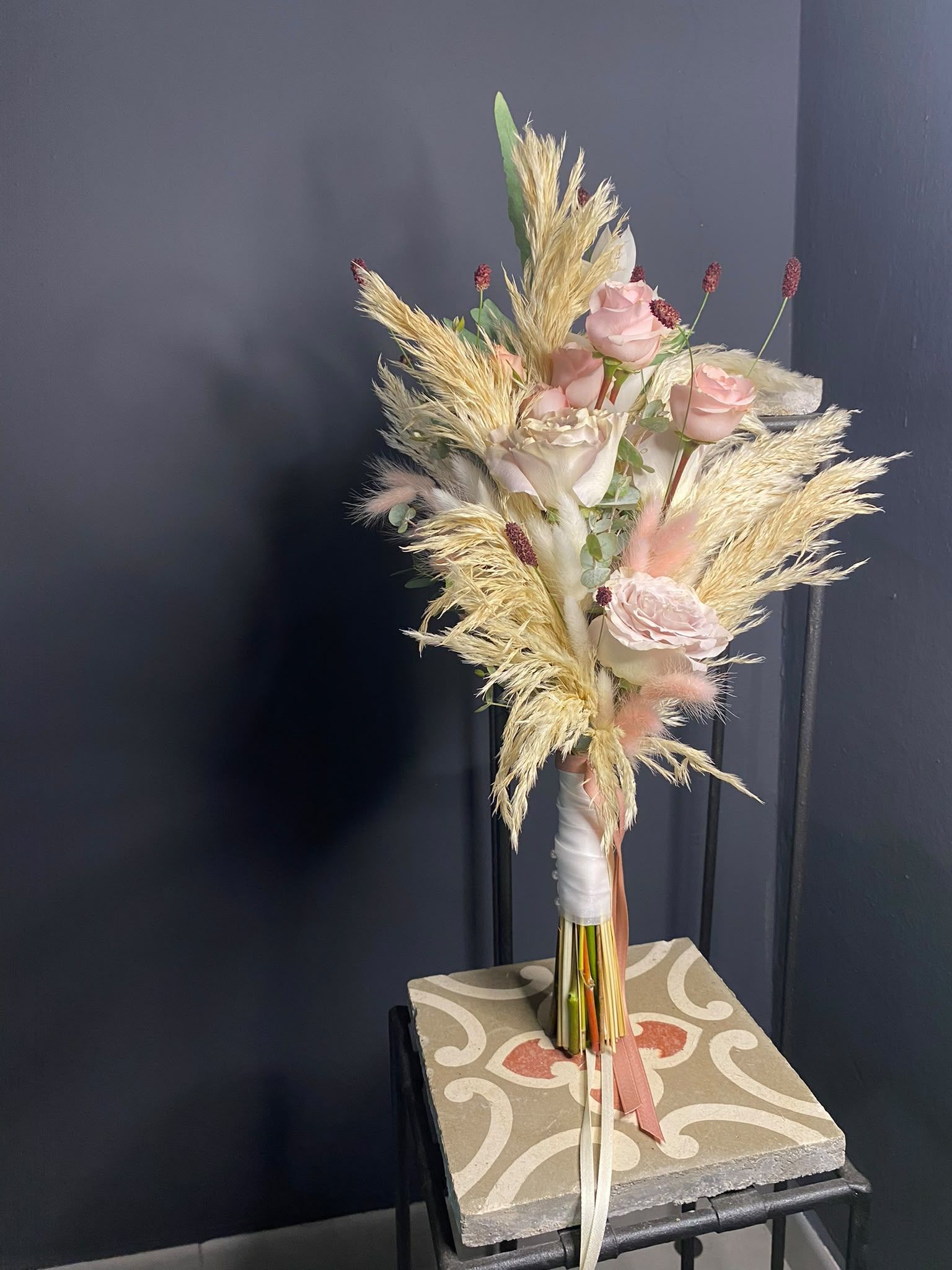 Arreglo floral seco en tonos rosa pastel y crema en un jarrón sobre un libro estampado contra una pared oscura.