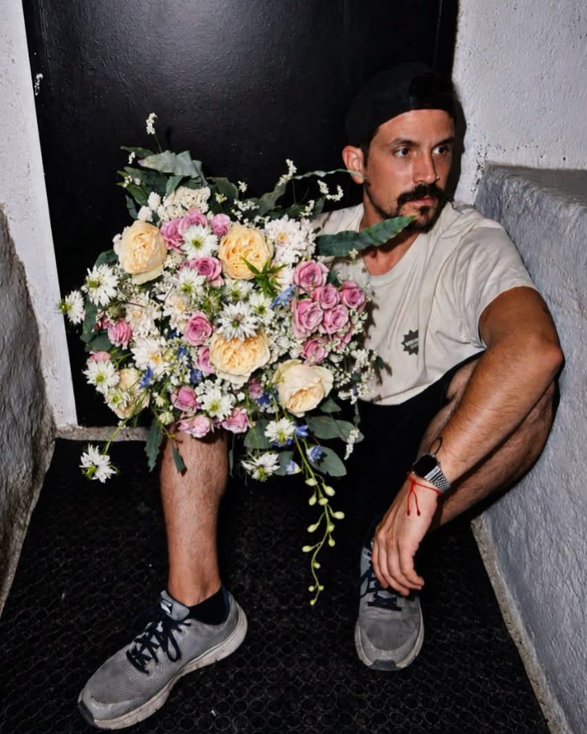 Un hombre sentado en el suelo sostiene un gran ramo de flores rosas y blancas, mirando hacia un lado.