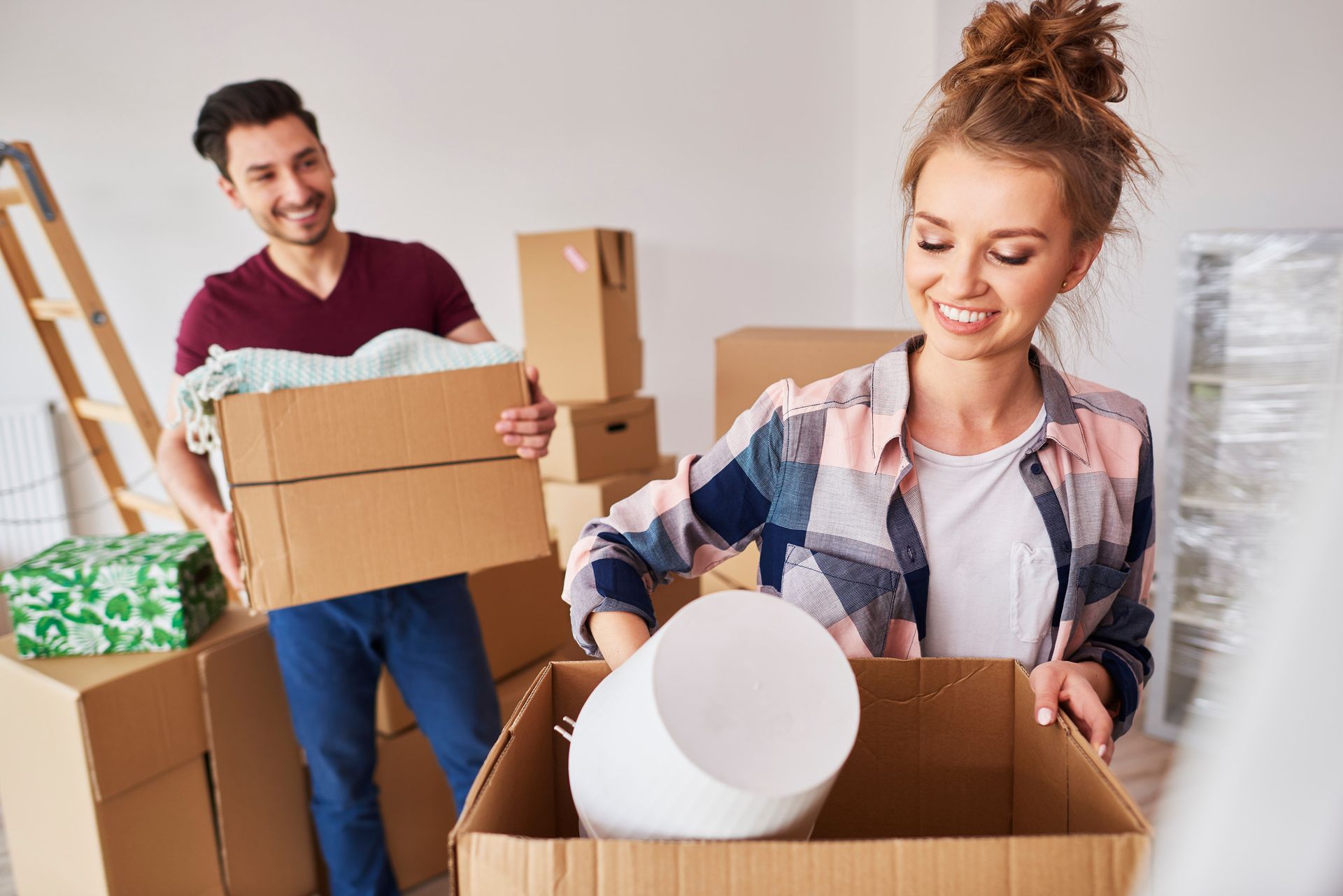 Pareja desempacando cajas en una nueva casa; el hombre sonríe sosteniendo una caja, la mujer desempaca un balde y sonríe.