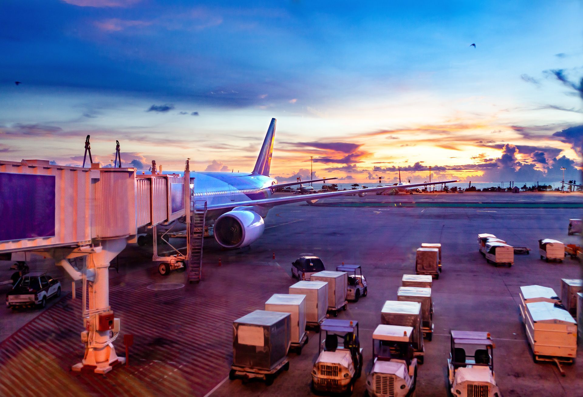 Avión en la puerta del aeropuerto durante la puesta de sol, con carros de equipaje y un cielo resplandeciente de colores.