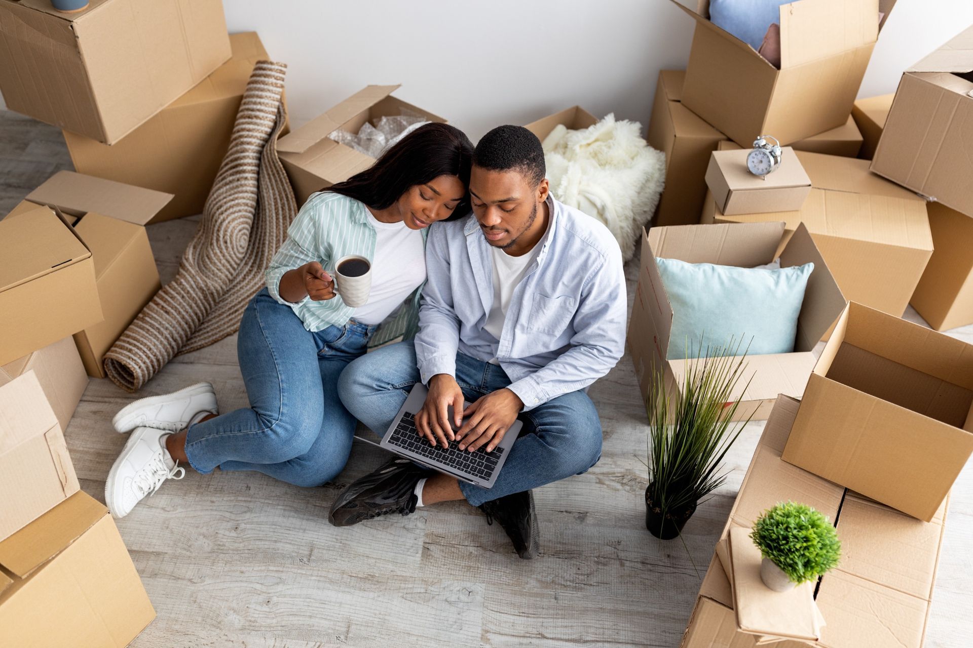 Una pareja sentada rodeada de cajas de mudanza: una mujer con café y un hombre con una laptop. Habitación luminosa, expresiones felices.