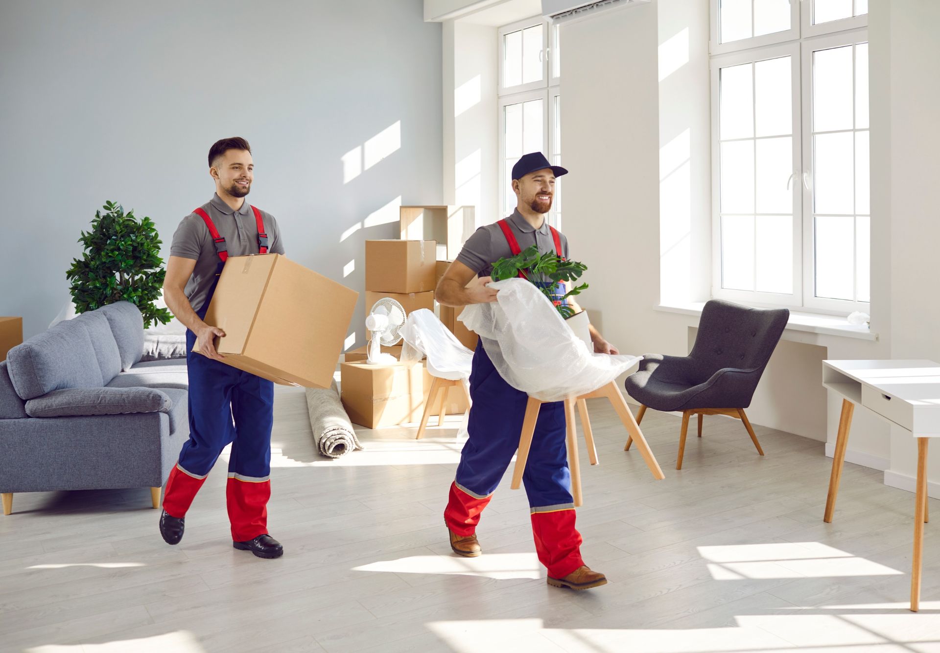 Dos trabajadores de mudanzas transportan artículos a una habitación vacía y luminosa con grandes ventanales.
