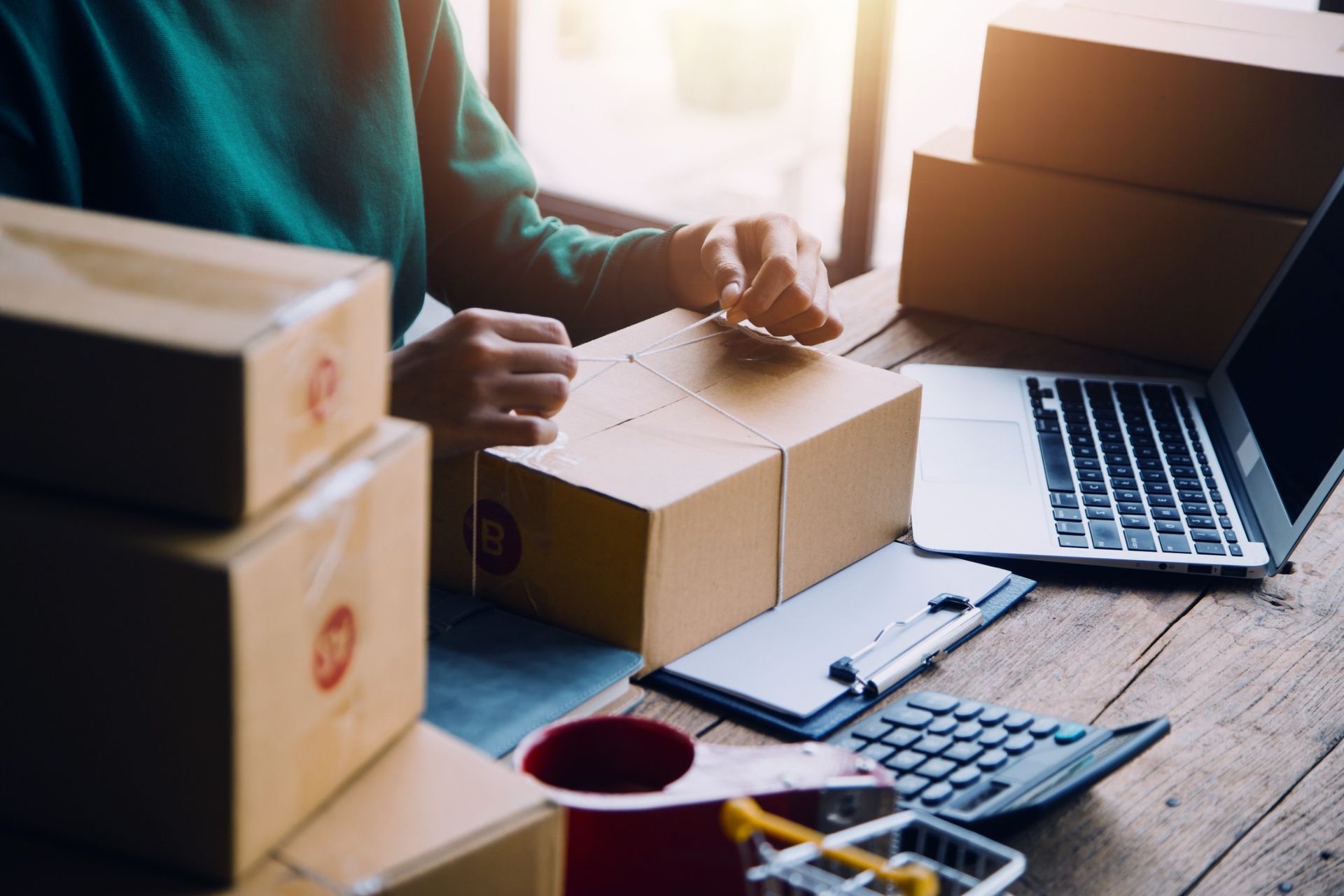 Persona empacando una caja, rodeada de paquetes, una computadora portátil, una calculadora y un pequeño carrito de compras.