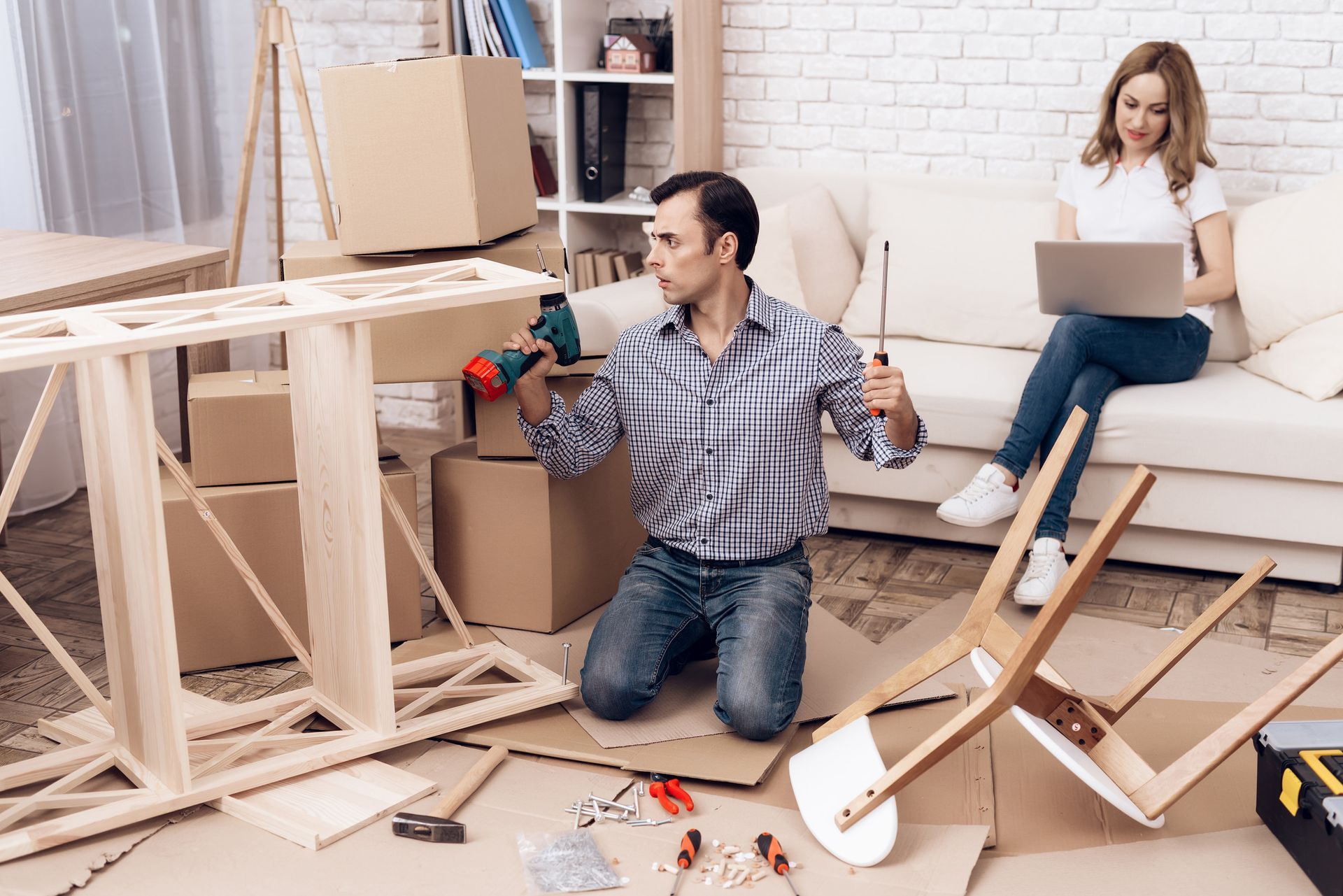 Hombre arrodillado, usando un taladro, ensamblando muebles; mujer en el sofá usando una computadora portátil en una sala de estar.