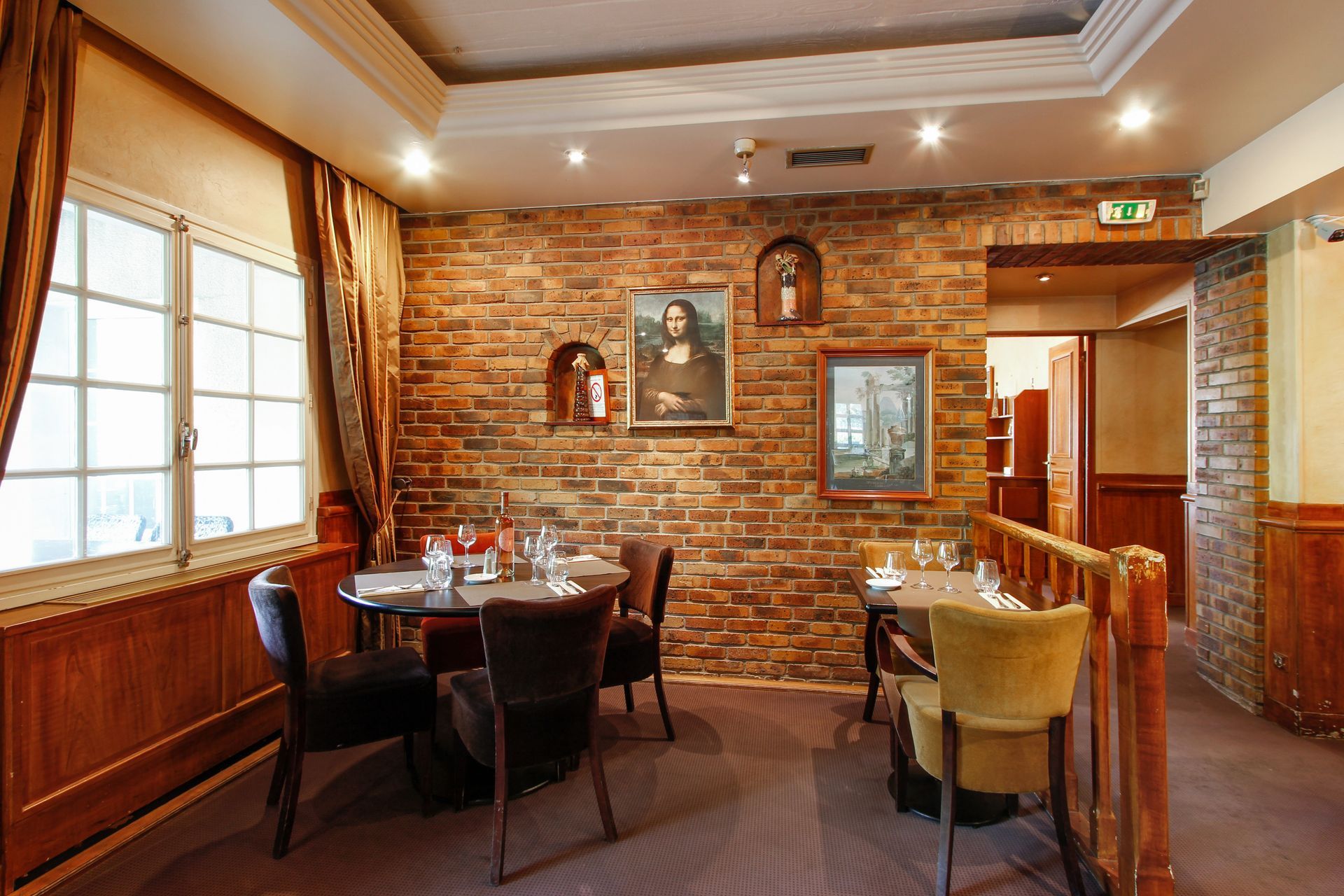 Une salle à manger de restaurant avec des tables, des chaises et des tableaux sur un mur de briques rustiques, éclairée par des spots encastrés.