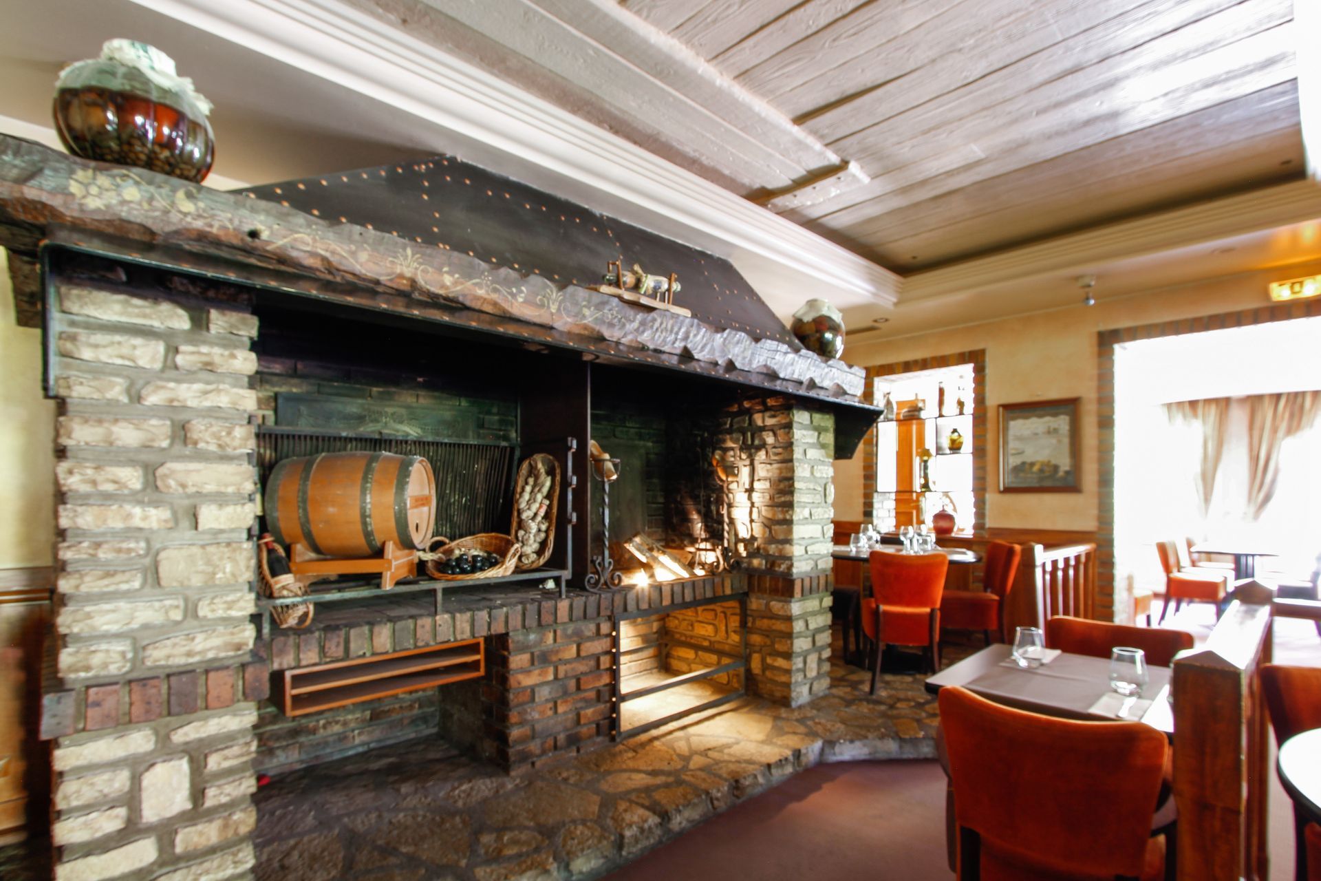 Intérieur de restaurant rustique avec une grande cheminée en briques, un tonneau en bois et des chaises aux tons chauds.