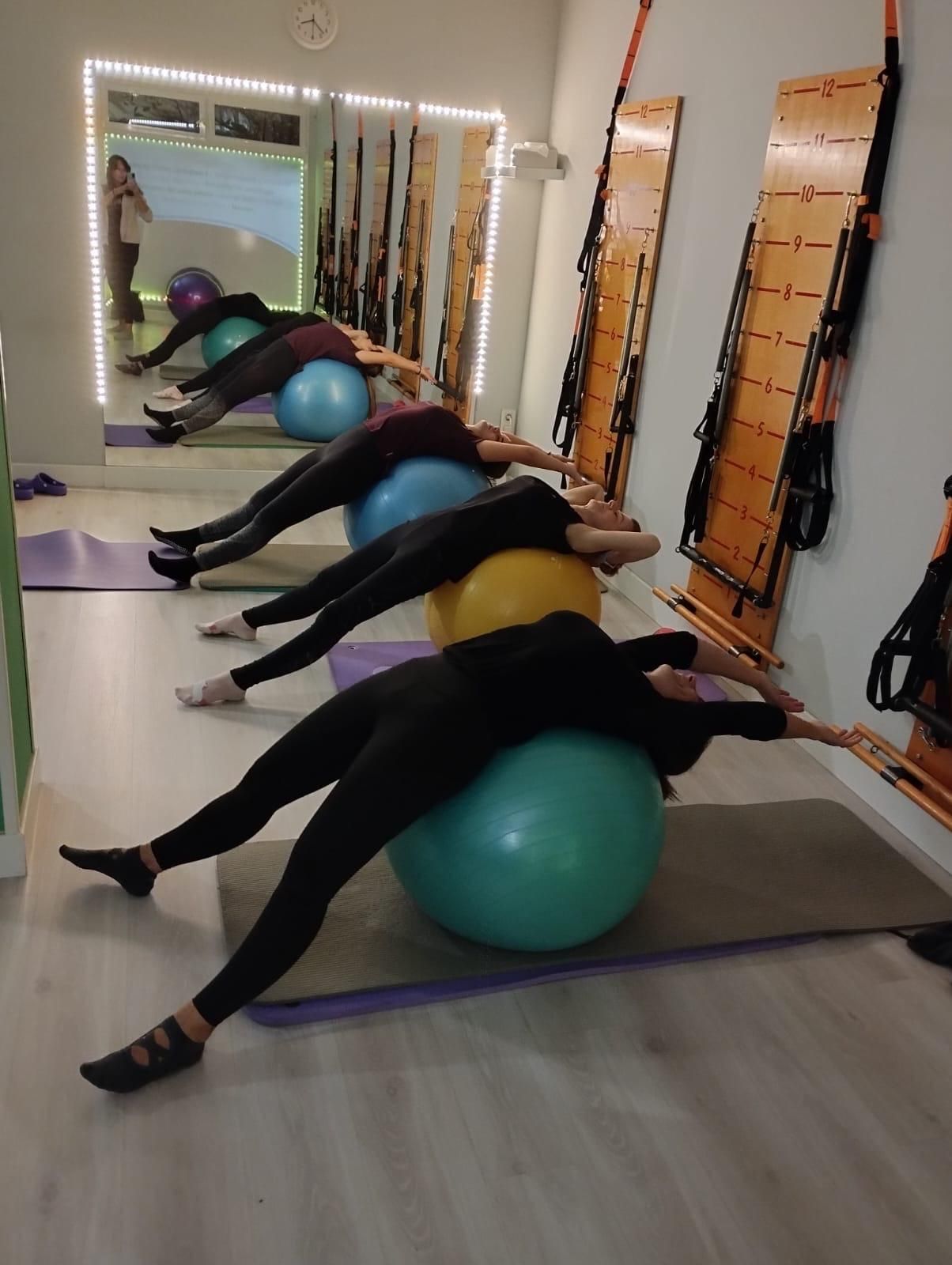 Personas haciendo ejercicio con pelotas grandes en un estudio con espejos y aparatos. Levantan los brazos.