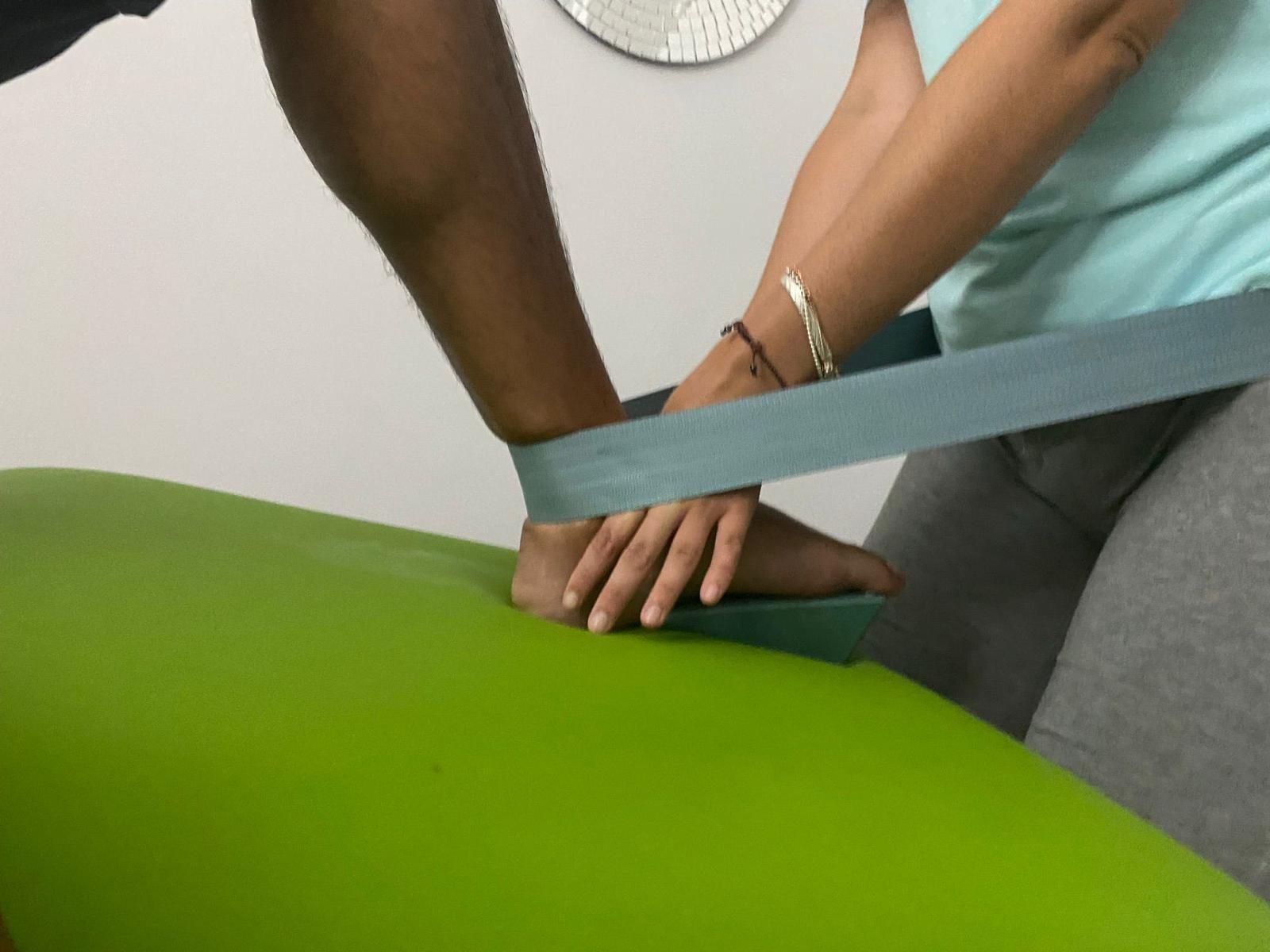 Persona estirando el tobillo de otra persona con una correa azul sobre una superficie verde.