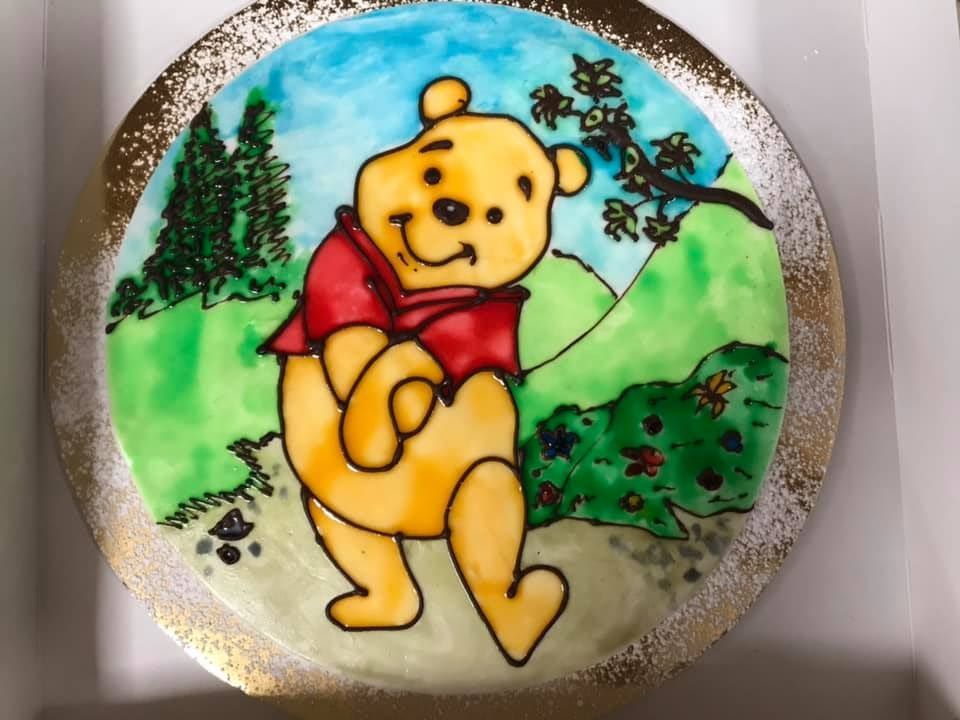Winnie the Pooh su una targa rotonda dipinta, in piedi in un verde paesaggio rurale con alberi e colline.