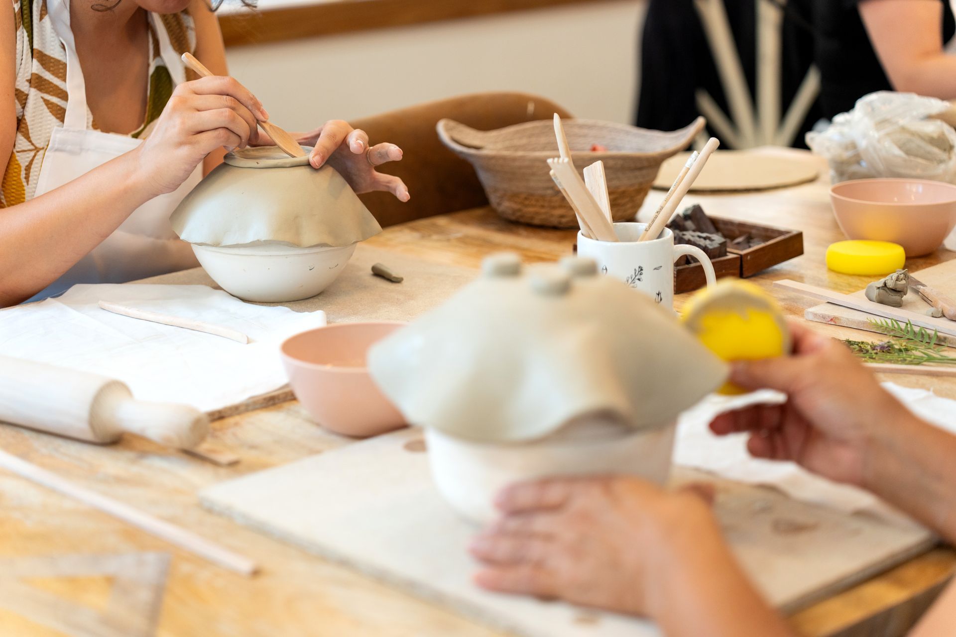 Des mains façonnent l'argile sur des tours de potier à une table d'atelier, entourées d'outils et de bols.